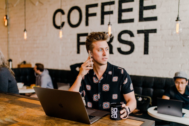 Young man talking on smartphone holding a mug of coffee infant of his laptop in a coffee shop